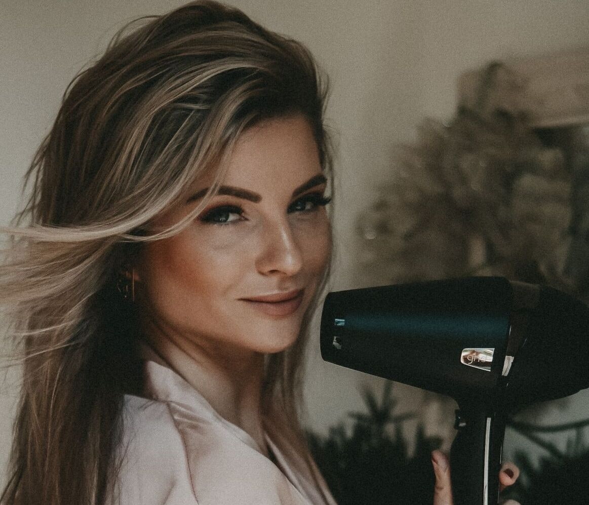 Haarpflegetipp richtiges Föhnen Frau mit braunen Haaren und blonden Strähnen hält einen Haarföhn in die Kamera