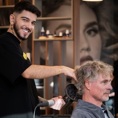 Styling Typberatung KLIPP Ein Friseurmitarbeiter blickt in die Kamera während er einem älteren Mann die Haare föhnt